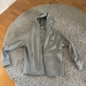 Polo Ralph lauren grå zip - Bra skick, använd fåtal gånger. Jag har tagit bort snören på den men skickar med de om man vill lägga tillbaka snörena. Inga fel på tröjan. Nypris 1600-2000. Säljs för att den inte används längre.