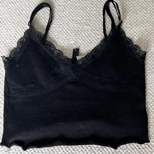 Svart linne från H&M Divided - Ett svart linne från H&M Divided med spetsdetaljer vid halsringningen och axelband. Linnet har en ribbad struktur. Bra skick. Längst ner på tröjan så snurrar den upp sig tyvärr men inte ett stort problem. Skriv för fler bilder eller frågor! 🖤