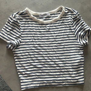 Randig topp från Zara - En vit och blå randig topp från Zara med korta ärmar. Toppen har en rund halsringning och är tillverkad i ett mjukt material.