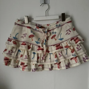 Vit kortkjol med svampmönster - En vit kortkjol med ett färgglatt svampmönster. Kjolen har volanger och ett knytband i midjan. Tillverkad av 65% polyester och 35% elastan.