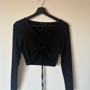 Svart topp med knytning framtill - Svart långärmad topp som man kan knyta framtill. Man kan vira banden runt kroppen och välja ifall man vill knyta en söt rosett antingen framtill eller på ryggen. Fin till kostymbyxor och fest! Storlek 36 och passar som S.