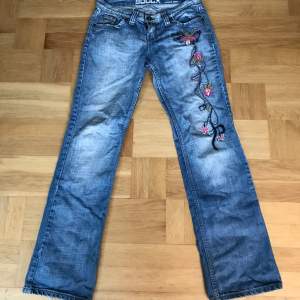 Lågmidjade bootcut jeans med super fina inbroderade blommor längst benet!! Midjemått rakt över= 42 cm Innerbenslängd = 85 cm Längden på hela byxorna= 108 cm 