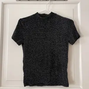 Glittrig svart t-shirt från Monki i storlek S. Turtleneck krage. Stretchigt material. Använd ett fåtal gånger, i fint skick. Bara att fråga om det är något du undrar! Kan mötas på Järntorget i Göteborg eller skicka, köparen står för frakt.