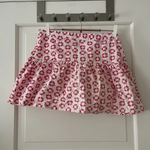 Kort kjol med blommor  - Kort volangkjol från X-Mail i storlek 40-42. Vit/mönstrad med rosa blommor. Knäpps i sidan med knappar. Knappt använd, i fint skick. Bara att fråga om det är något du undrar! Kan mötas på Järntorget i Göteborg eller skicka, köparen står för frakt.