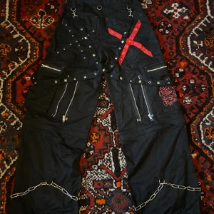 emo/goth byxor - tripp nyc liknande vida byxor med röda detaljer, nitar, kedjor och dragkedjor. avtagbar nedre del så de kan bli till längre cargoshorts! skicket är mycket bra utöver en liten nit som saknas. 