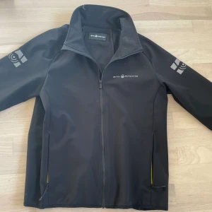 Sail Racing Soft Shell jacka, 170 - Svart jacka från Sail Racing. Märket är i reflex och sitte på bröstet, ryggen och ärmarna. Inköpt våren-23, är i fint skick. Nypris 1499:-