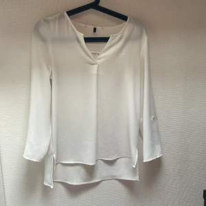 Vit blus - En elegant vit blus med långärmad design. Blusen har en enkel och stilren look med en lätt v-ringad hals och en lös passform.