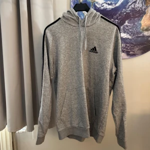 Grå hoodie från Adidas - Säljer en snygg och bekväm grå hoodie från Adidas. Den har svarta ränder längs ärmarna och Adidas-loggan på bröstet. Perfekt för både träning och vardag. Hoodien har en känguruficka och dragsko i huvan.