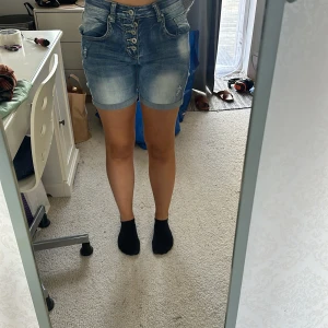Blå jeansshorts från Place du Jour - Säljer ett par snygga blå jeansshorts från Place du Jour i storlek 36. De är högmidjade och har en cool knappdetalj framtill. Perfekta för sommaren! Shortsen är i nyskick och har en normal passform. Materialet är mestadels bomull med lite elastan för extra komfort.