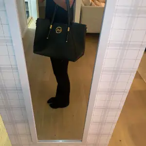 Säljer en snygg svart handväska från Michael Kors. Väskan är i skinn och har två handtag. Den har en guldig MK-logga på framsidan och gulddetaljer vid handtagen. Perfekt för både vardag och fest!