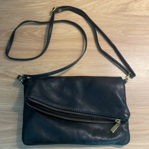 Svart axelväska med dragkedja - En svart axelväska i skinn med en justerbar axelrem. Väskan har en dragkedja på framsidan och en dragkedja för huvudfacket. Insidan är fodrad med svart tyg och har en liten innerficka med dragkedja.