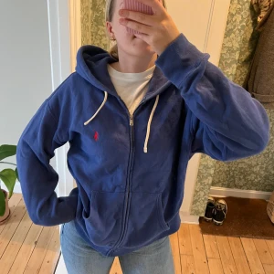 Blå hoodie från Ralph Lauren - Säljer en blå hoodie från Ralph Lauren. Den har en dragkedja framtill och en liten röd logga på bröstet. Hoodien har långa ärmar och en justerbar huva med vita snören. Perfekt för en avslappnad stil!