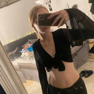 Svart knytblus med transparenta ärmar - Säljer en snygg svart knytblus med transparenta långa ärmar. Blusen är croppad och har en knytning framtill som ger en fin detalj. Perfekt för fest eller en utekväll! 🖤