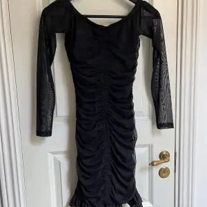 Snygg svart klänning med långa genomskinliga ärmar och en volangdetalj i nederkanten. Klänningen är figursydd.