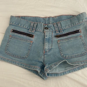 Blå jeansshorts med dragkedjor - Ett par blå jeansshorts från levis med två framfickor och dragkedjor. Shortsen har en knapp och dragkedja framtill samt en bakficka. Det finns även en dekorativ snörning på baksidan. Lågmidjade! Midjemåttet rakt över är ca 39cm och längden från midjan till grenen är ca 21cm💕Använd gärna köp nu