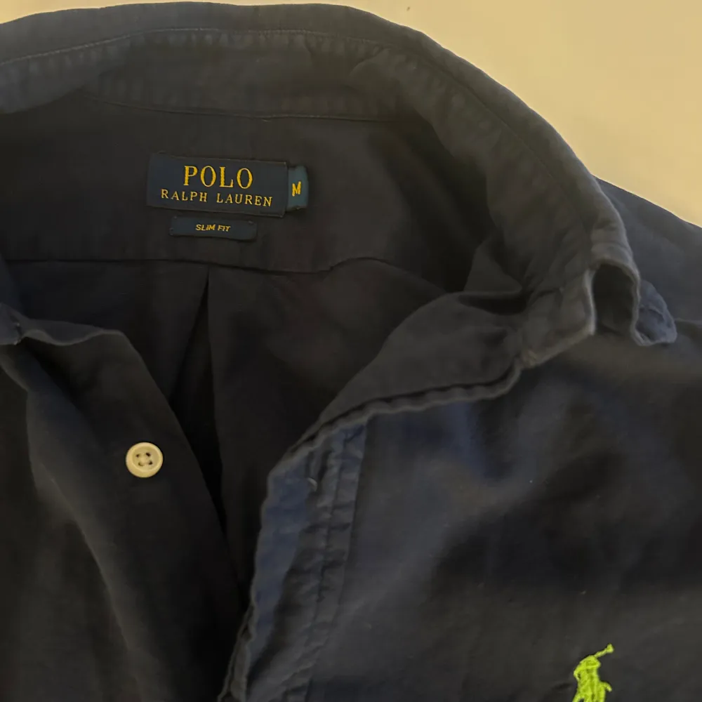 En mörkblå skjorta från Polo Ralph Lauren i storlek M. Skjortan har långa ärmar och knappar framtill. Den är av slim fit-modell och har en broderad grön logotyp på bröstet.. Skjortor.