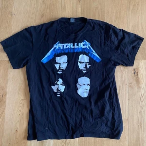 Svart Metallica t-shirt från Rock Yeah - En svart t-shirt från Rock Yeah med Metallica-tryck. T-shirten har korta ärmar och ett tryck med bandmedlemmarnas ansikten på framsidan samt Metallica-logotypen i blått på både fram- och baksidan.