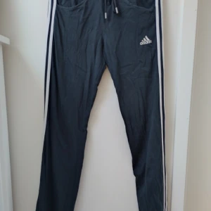 Svarta träningsbyxor från Adidas - Ett par svarta byxor från Adidas med vita ränder längs sidorna. Byxorna har snöre i midjan för justerbar passform och två fickor framtill.