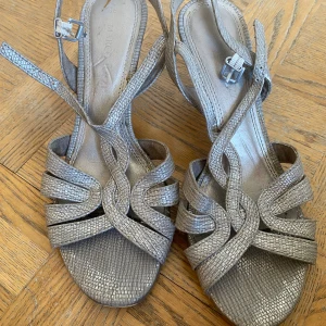 Silvriga sandaler med klack - Ett par eleganta silvriga/guldiga sandaler med klack. Sandalerna har ett flätat mönster på remmarna och ett justerbart spänne runt ankeln. De är i storlek 38 och har en bekväm klackhöjd.