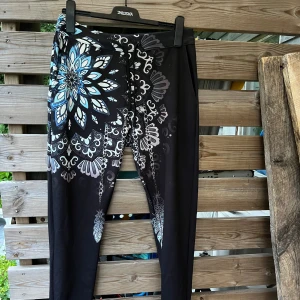Coola baggy mjukisbyxor i nyskick! - Super snygga mjukisbyxor med stor mandala/blomma, fjädrar. Endast provade men aldrig tvättade. Mjuk insida i bomull, utsida aningen skimrande polyester. 