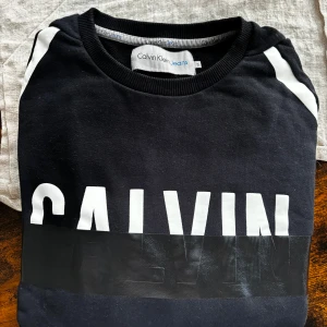 Calvin Klein tjocktröja - Tjocktröja från Calvin Klein i cool svart-vit design och stor logga framtill.  Har två smidiga fickor vid magen. 