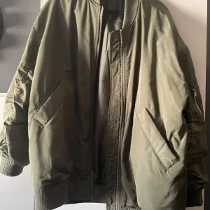 Grön bomberjacka - En grön bomberjacka med dragkedja och ribbade muddar vid ärmslut och nederkant. Jackan har två stora fickor framtill och en mindre ficka med dragkedja på ärmen. Passar mig även att jag är en L/XL, storlek XS på jackan. Finns på flera sidor. 
