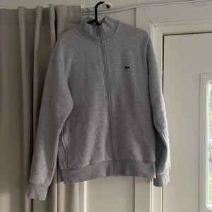 Grå Lacoste sweatshirt med dragkedja, strl M - Lacoste tjocktröja med dragkedja (grå, strl M).Ny pris är som på bilden 1 599kr, köpt för ganska exakt 3 månader sen men endast använda ett fåtal gånger och den används inte längre. Den är nyskick Inge fläckar eller skador överhuvudtaget.