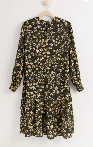 Klänning - Mönstrad klänning i nästintill oanvänt skick  En mönstrad klänning med midilängd, volang på kjolen och smock vid ärmslut. Plagget är gjort i en återvunnen polyesterblandning. 