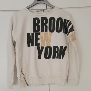 Sweatshirt med tryck - En vit sweatshirt med tryck "brooklyn new york". Har en dragkedja som detalj längst ner på ena sidan. Har 2 fläckar på bokstäverna i fram, men märks inte supertydligt. 52 cm från hals till midjeslut. Armlängd från armhåla är ca. 45 cm. 