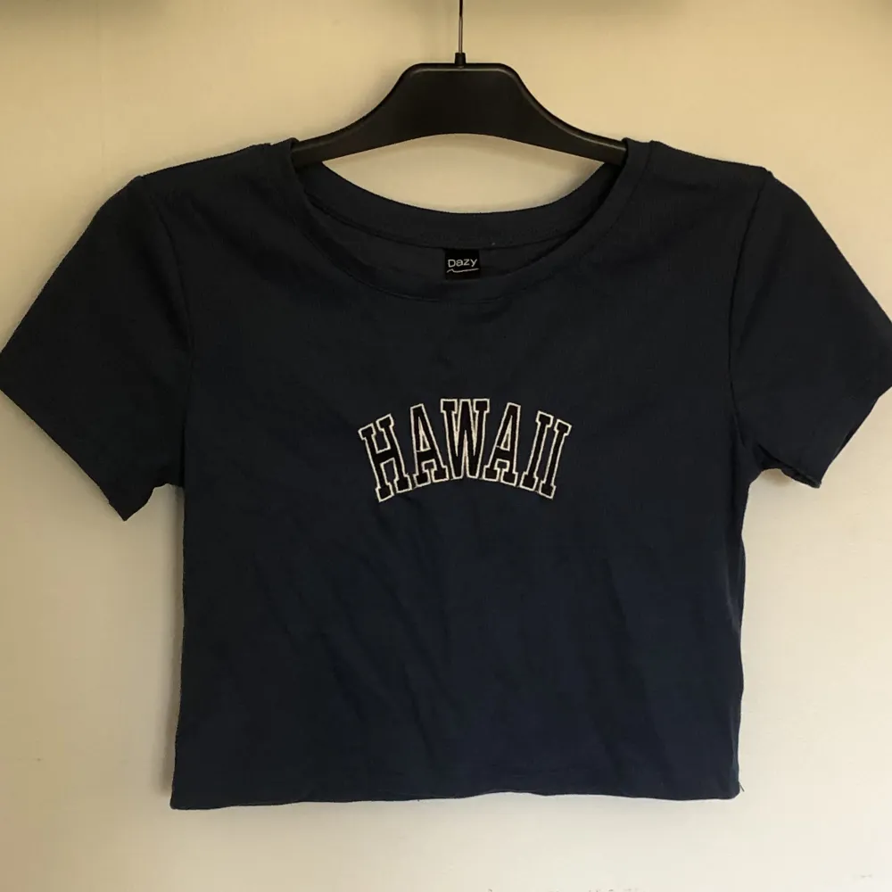 Detta är en croppad T shirt med texten Hawaii. Denna är i stl L och är mörk blå, även om det inte kommer fram så bra i bilderna. Toppen är endast använd ett fåtal gånger och är i fint skick.. Toppar.