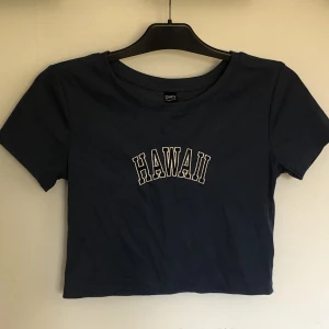 Croppad T shirt i stl L. - Detta är en croppad T shirt med texten Hawaii. Denna är i stl L och är mörk blå, även om det inte kommer fram så bra i bilderna. Toppen är endast använd ett fåtal gånger och är i fint skick.