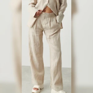Linnebyxor  - Nya  Linnebyxor med lös passform i strl M. Linnebyxorna är beigea och har veck framtill. De har raka ben, resår bak i midjan, bälteshällor, sidofickor och en passpolerad ficka bak