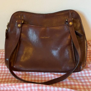 Brun läderhandväska - En snygg och praktisk handväska i äkta läder! Den är i använt men fräscht skick, den har en defekt - en vattenskada på baksidan som visas på den andra bilden. Många fack och fickor på insidan och utsidan!! Skriv för närmare bilder/mått m.m.💓