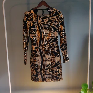 Klänning  - Svart/guldig paljettklänning med djup uringad rygg och långa ärmar som passar till fest. Svart tyg under så det inte kliar av paljetterna. 