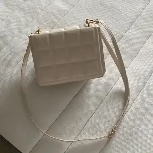 En handväska i färgen vit/beige med gulddetaljer. Axelbandet är justerbart och går att fästa vid både sidorna och på väskan. 