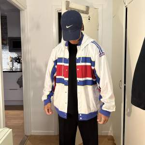 En riktigt snygg vintage track jacket från adidas med avtagbar luva. Tunn och skön. Den är hel utan några större märken, på en av knapparna har lite färg lossnat.. storlek L men passar M skulle jag säga. Är 180cm 72kg.  Vit röd och blå.