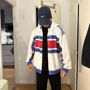 Adidas vintage jacka - En riktigt snygg vintage track jacket från adidas med avtagbar luva. Tunn och skön. Den är hel utan några större märken, på en av knapparna har lite färg lossnat.. storlek L men passar M skulle jag säga. Är 180cm 72kg.  Vit röd och blå.
