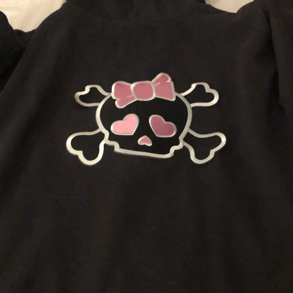 En tjock tröja med dragkedja. Den har en döskalle med en rosa rosett på ryggen, döskallen håller på och lossna lite på hörnen.. Hoodies.