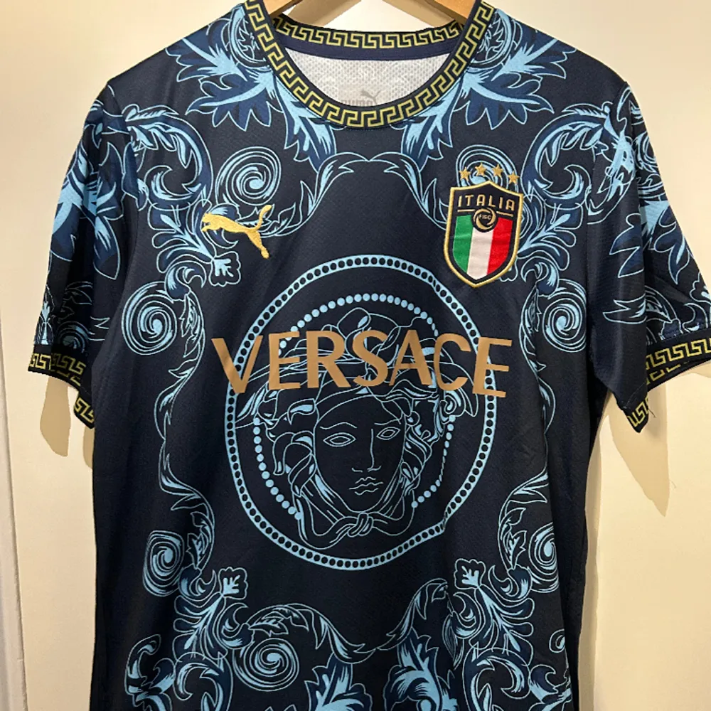 Mörkblå Italien X Versace T-shirt med Versace loggor runtom och guldiga detaljer samt text. Helt ny med etikett i storlek M.. T-shirts.