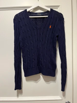 Marinblå kabelstickad tröja  - En elegant Ralph Lauren POLO tröja med en v urringning. I mycket bra skick och har används ett fåtal gånger. Det är 100% bomull i materialet och är väldigt skön att ha på sig med eller utan Också skjorta. 
