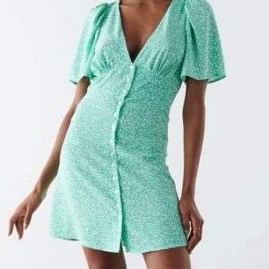 Klänning  - Miniklänning med korta ärmar. Klänningen är grön och har en lätt vävd kvalitet. Den har v-ringning, knappstängning framtill och rynkningar på bröstet och på axlarna.