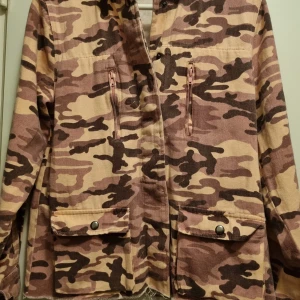 Kamouflage jacka  - Rosa kamouflage mönstrad skjortjacka med knappar för knäppning. Två bröstfickor med dragkedja och två fickor fram nedtill med knappar 