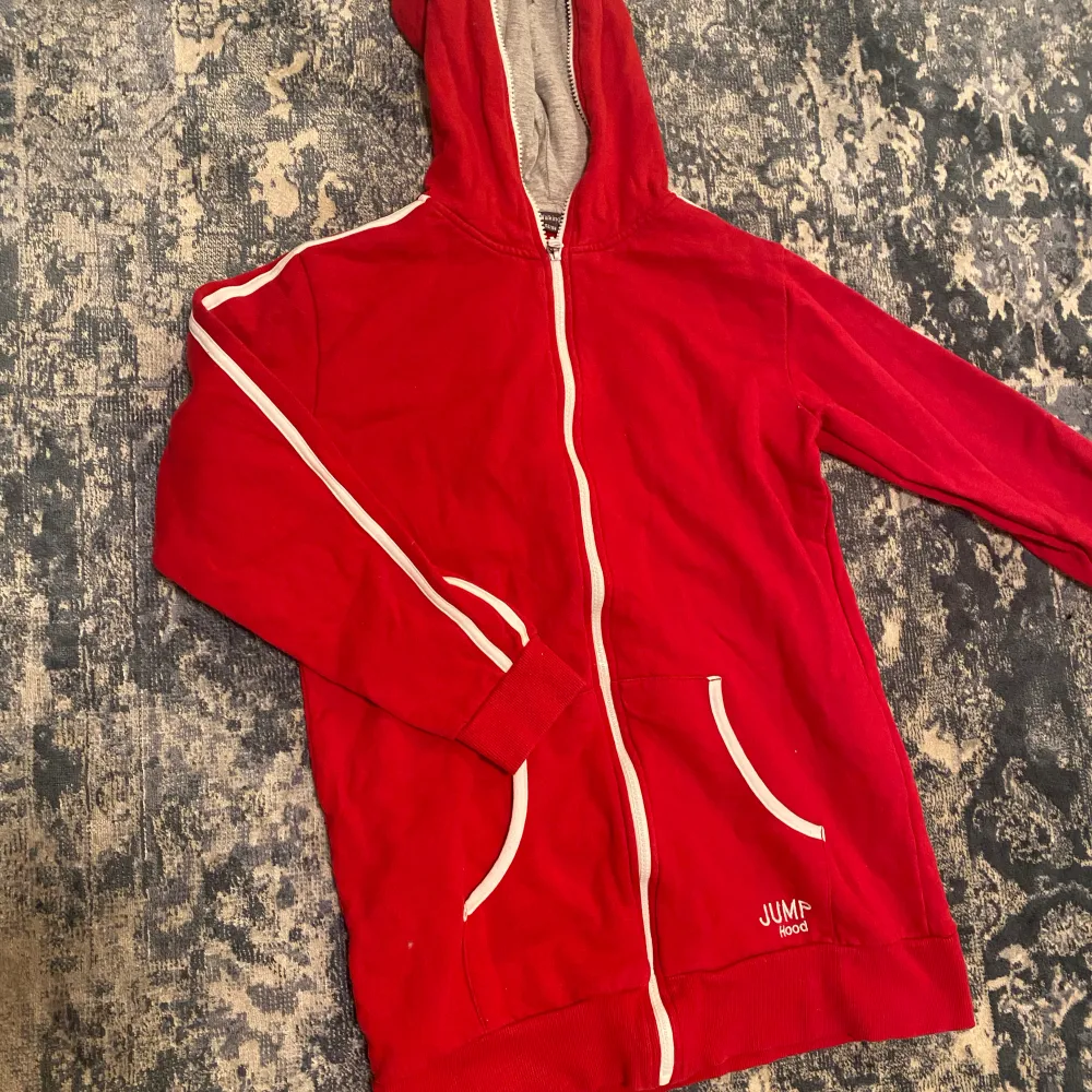 En röd zip up hoodie med vita detaljer, lite kortare model i armarna, text på ryggen. Hoodies.