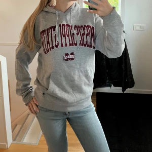 Grå Mississippi State hoodie - Säljer en grå hoodie med tryck från Mississippi State i vinrött och vitt. Tröjan har huva med snörning och en stor magficka. Perfekt för chill dagar eller när du vill ha en sportig look.