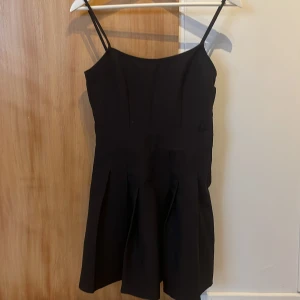 Svart kort klänning från H&M Divided - Svart klänning med smala axelband från H&M Divided. Modellen är kort med veckad kjol och har en diskret dragkedja i sidan. Stilren och enkel design som ger en snygg siluett. Perfekt för dig som gillar minimalistisk stil.