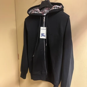 Svart Burberry hoodie med dragkedja - Svart hoodie från Burberry med klassiskt rutigt foder i huvan och broderad logga på bröstet. Tröjan har dragkedja framtill, snörning vid huvan och långa ärmar. Perfekt för dig som gillar stilrena och lyxiga streetwear-plagg.