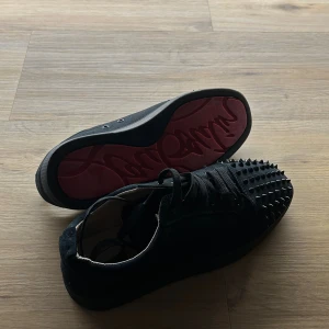 Christian Louboutin svarta sneakers med nitar - Svarta sneakers från Christian Louboutin med coola svarta nitar på tån och klassisk röd sula. Skorna har snörning och är tillverkade i mocka med skinnfoder. Perfekt för dig som vill sticka ut med en edgy och lyxig stil.