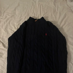Ralph Lauren half zip - Säljer en mörkblå stickad tröja från Polo Ralph Lauren med klassiskt kabelstickat mönster och röd logga på bröstet. Tröjan har half zip vid halsen och ribbade muddar. Perfekt för dig som gillar stilren och tidlös design.