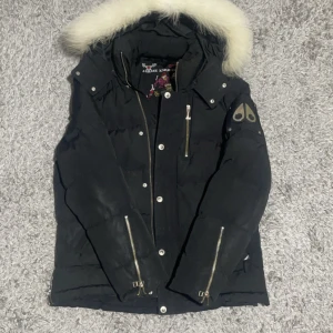 Svart dunjacka Moose Knuckles med päls - Svart dunjacka från Moose Knuckles med fluffig vit päls på huvan. Jackan har flera dragkedjor, knappar och coola detaljer på ärmen. Insidan har snyggt tryck och logga. Perfekt för kalla dagar och har en riktigt schysst streetstil.