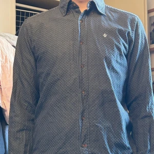 Mörkblå prickig skjorta med krage - Snygg mörkblå skjorta med små vita prickar och klassisk krage. Skjortan har långa ärmar, knappar framtill och en liten broderad logga på bröstet. Perfekt för dig som gillar stilrena detaljer och mönster som sticker ut lite lagom.